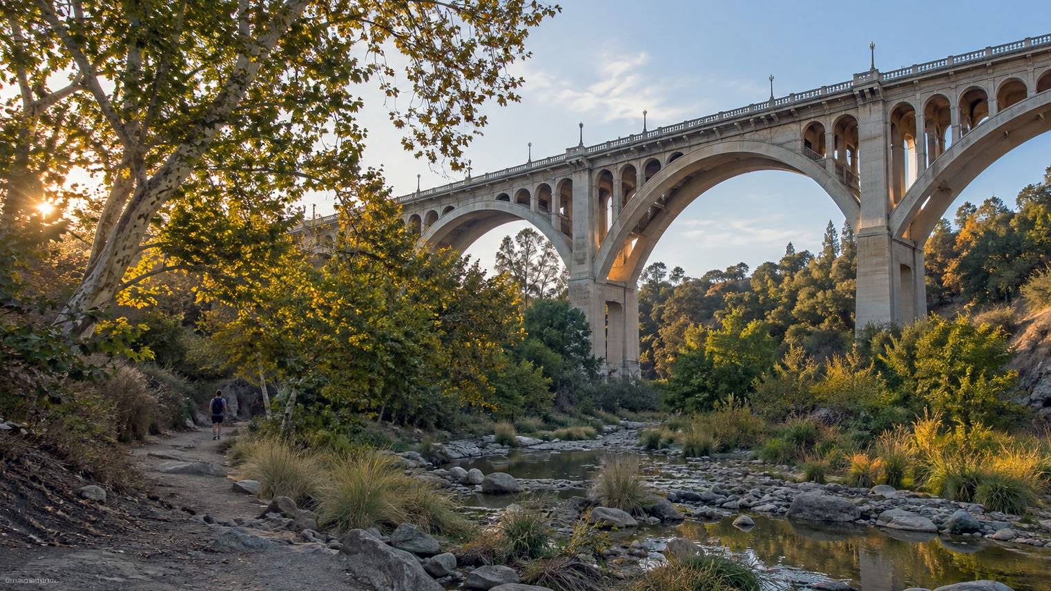 A concrete arch bridge high above the Arroyo Seco, a hiker on the streambed trail among sycamores and boulders