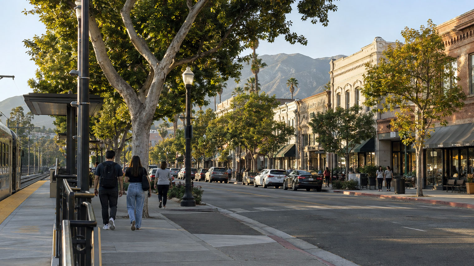 A couple walking a tree lined Old Town sidewalk beside a light rail platform, storefronts and mountains ahead