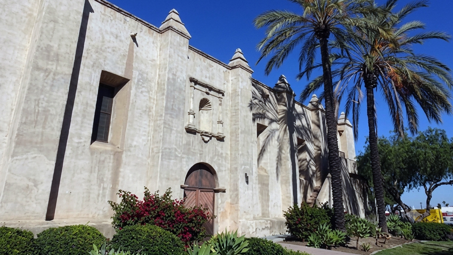 Mission San Gabriel Arcangel fortress wall, capped buttresses and arched wooden door behind bougainvillea and palms.