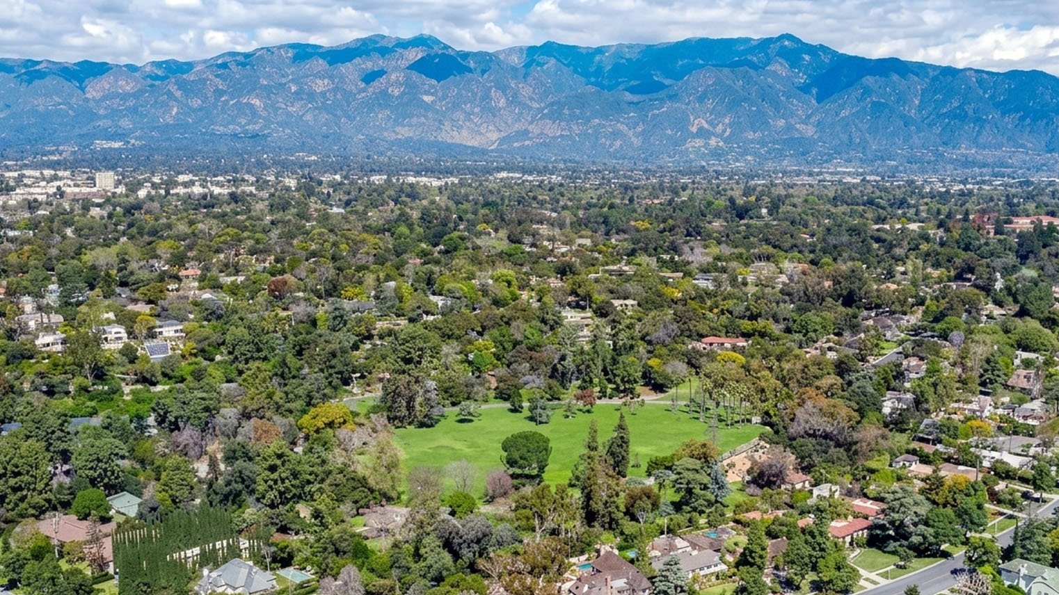 Aerial view of San Marino, a broad green park amid tree covered blocks, the San Gabriel Mountains across the horizon.