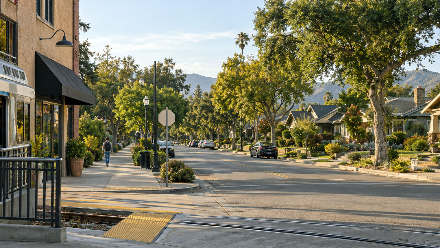 A rail crossing at a corner storefront, with Craftsman bungalows down a tree lined street and mountains beyond